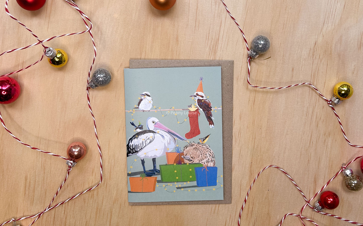 Greeting card with a festive family Christmas celebration with unique charm of Australian wildlife starring Kookaburras, a Pelican, Echidna, Blue Wren Spotted Pardalote, and a tiny sugar glider all decked out in holiday cheer.