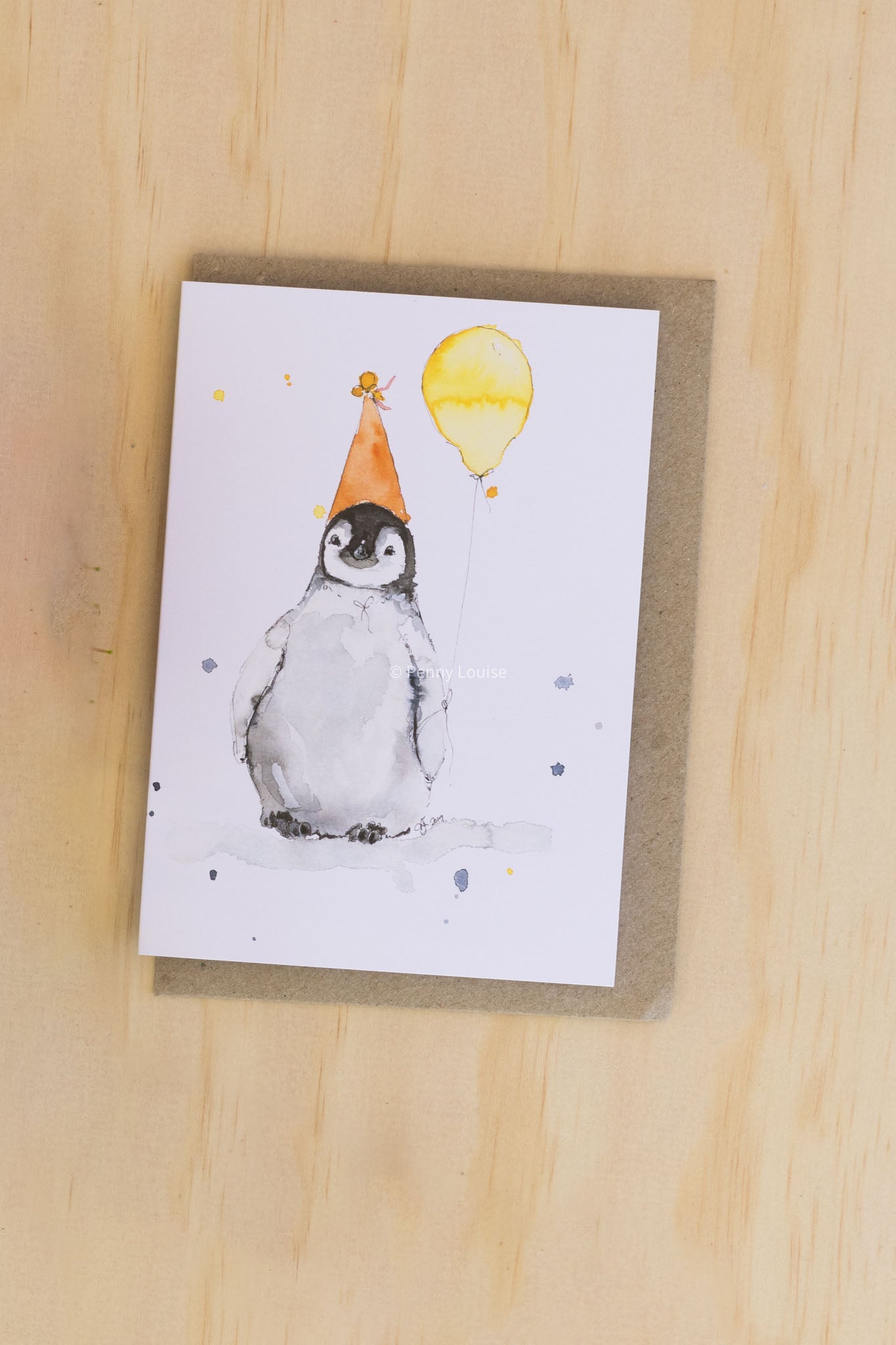 Greeting card with a penguin wearing a party hat and yellow balloon on a wooden surface