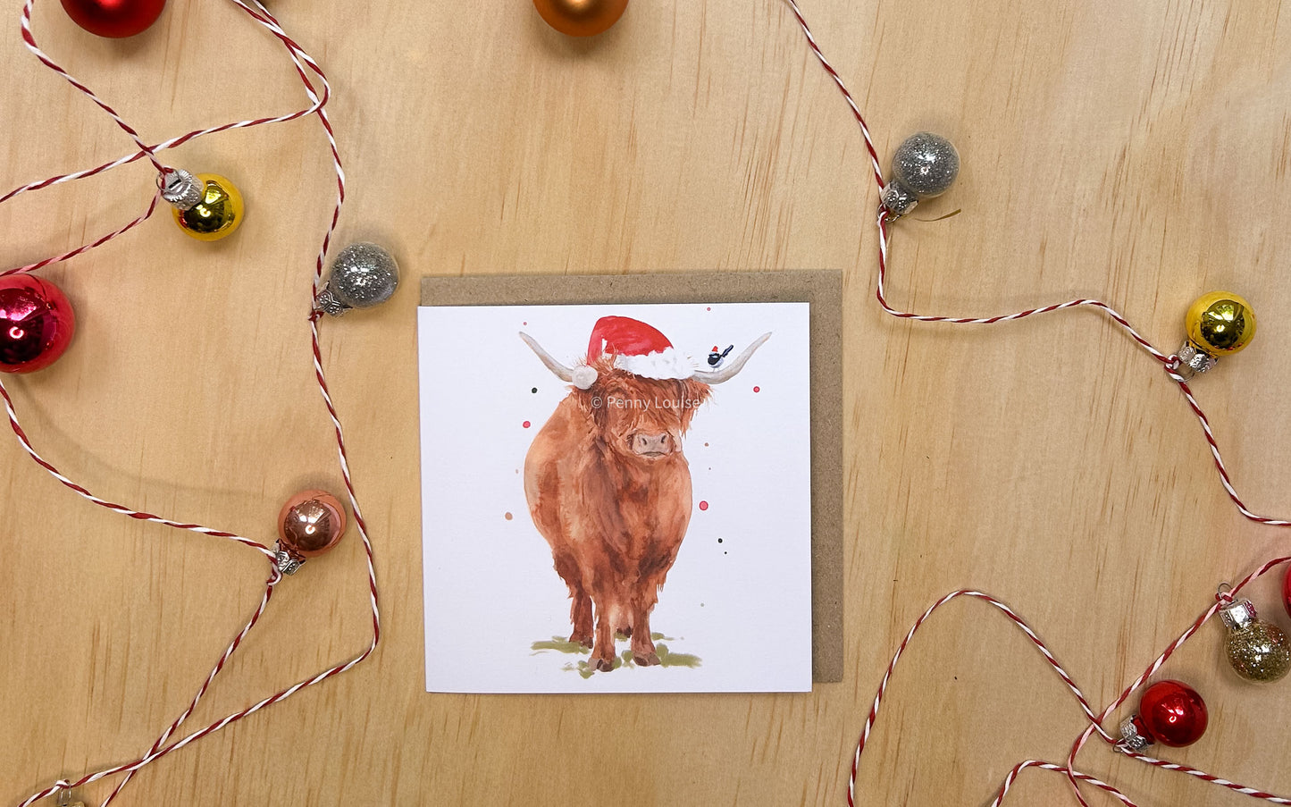 Card with a highland cow named Hilda wearing a Santa hat on a wooden surface with string lights.